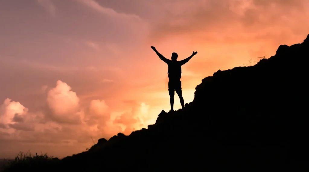 Person standing on a mountain with arms raised at sunrise.